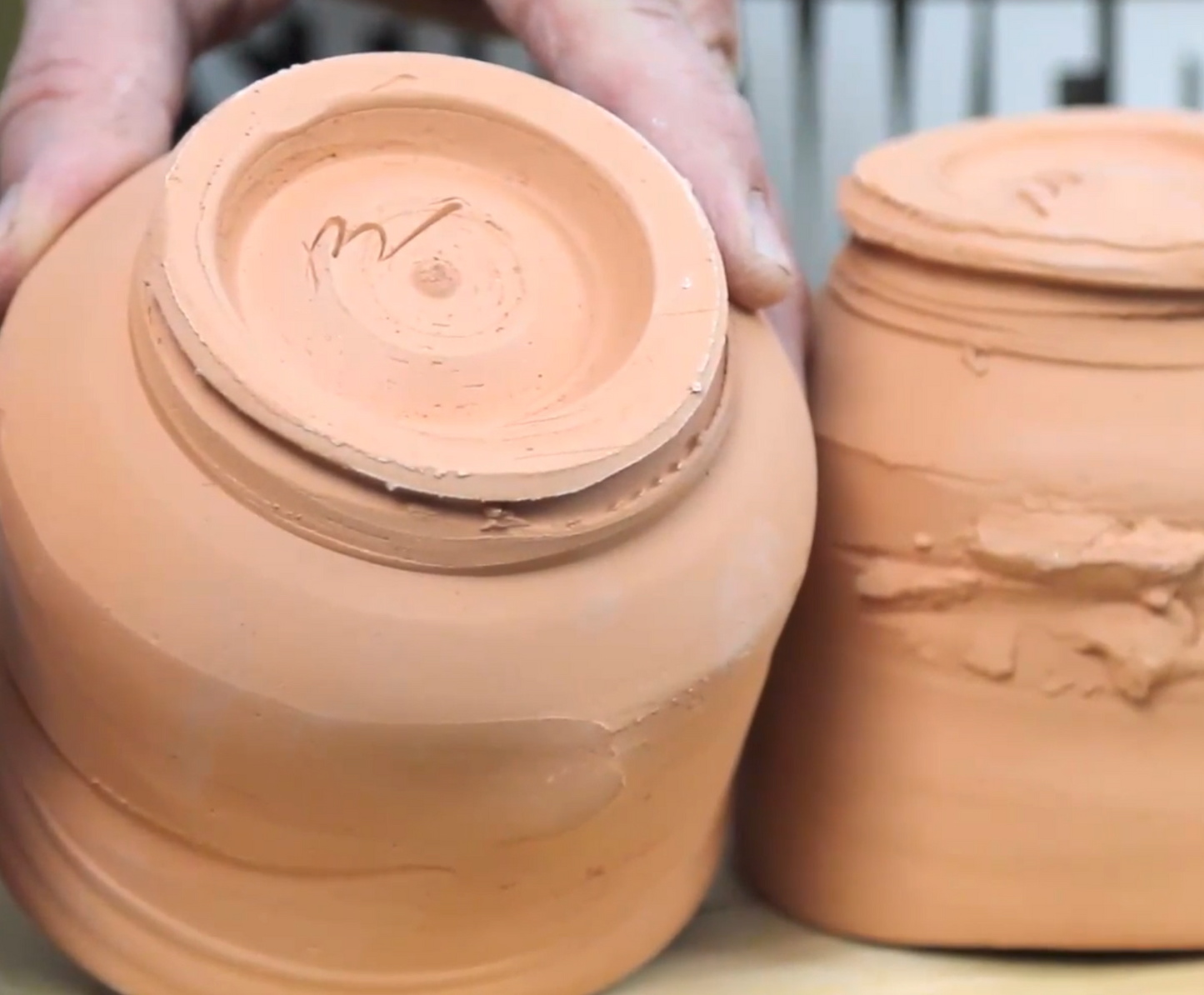 Two terracotta pots, one being held by a hand, on a neutral background.