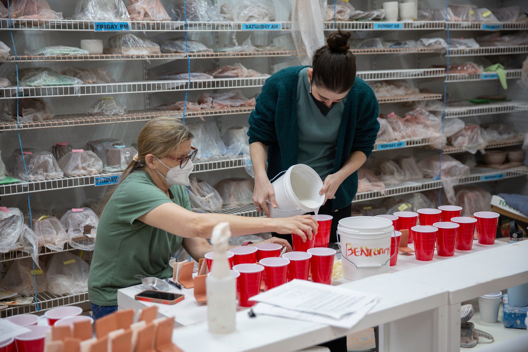 Two people in green shirts in a ceramics studio setting with shelves and red cups on a table.