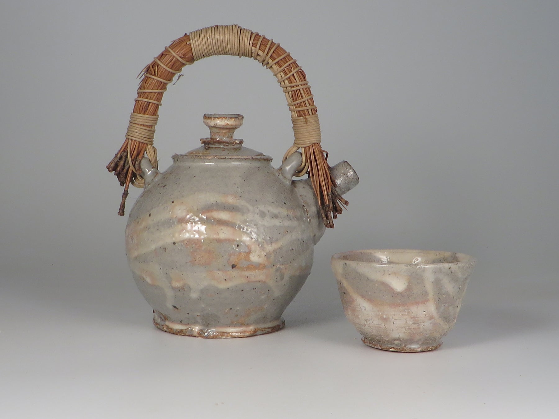 Ceramic teapot with woven handle and matching cup on a gray background