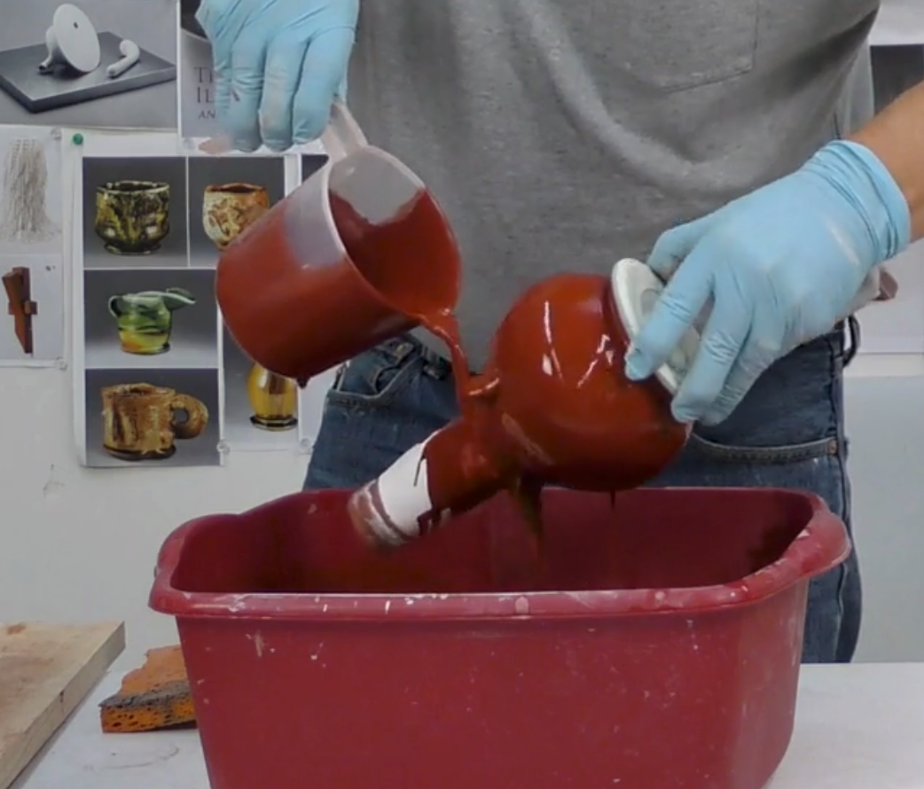 Artist John Britt's gloved hands pouring red liquid from a ceramic container into a bucket, with a display of ceramic items in the background.